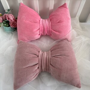 Peut inclure: Deux coussins décoratifs en forme de nœud. Le coussin du haut est en velours rose et le coussin du bas est en velours rose foncé.