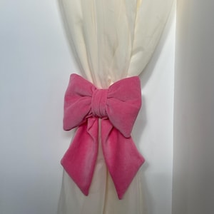 May include: A pink velvet bow tie curtain tieback. The tieback is tied around a pair of white curtains.