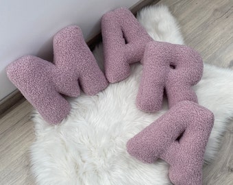 Könnte beinhalten: Vier Plüschbuchstaben, die den Namen "MARA" in einem hellen Rosa buchstabieren. Die Buchstaben bestehen aus einem weichen, flauschigen Material und sind auf einem weißen, flauschigen Teppich angeordnet.