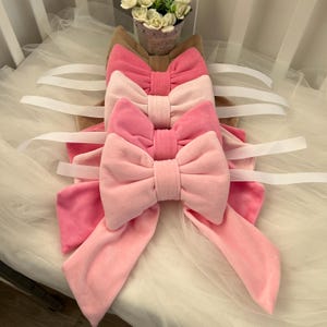 May include: A set of four pink velvet bows with white satin ribbon ties. The bows are arranged in a stack, with the largest bow on the bottom and the smallest bow on top.