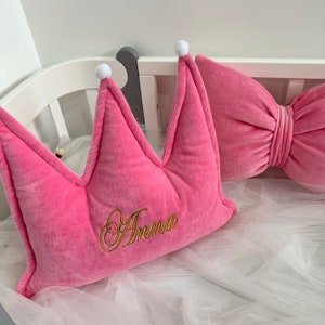 以下が含まれることがあります： 白いポンポンと金糸で「Anna」と刺繍されたピンクのベルベット製の王冠の形をした枕