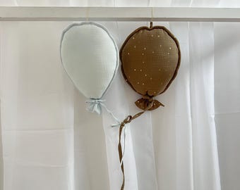 Peut inclure: Deux décorations de ballons en tissu suspendues à une tringle blanche. Un ballon est bleu clair et l'autre est brun avec des points dorés.