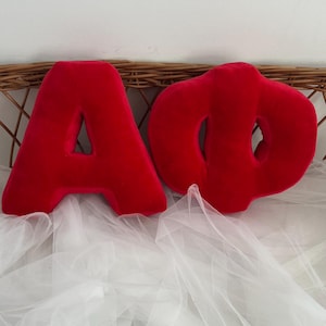 Puede incluir: Se muestran dos letras rojas de felpa, una "A" y una "O". Las letras están hechas de un material suave y aterciopelado. Descansan sobre una tela blanca y transparente, con una cesta tejida en el fondo.