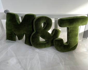 May include: Three large, plush letters spelling "M&J" in a dark green color. The letters are sitting on a white tulle fabric.