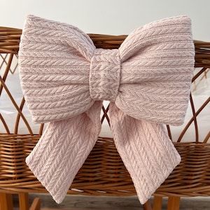 May include: A large pink knitted bow with a cable knit pattern. The bow is tied with long tails and is hanging on a wicker chair.