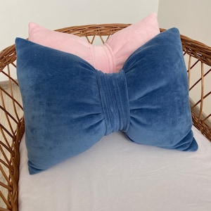 Peut inclure: Deux coussins décoratifs en velours dans un panier en osier. Le coussin du dessus est en velours rose et le coussin du dessous est en velours bleu. Les deux coussins ont la forme d'un nœud.