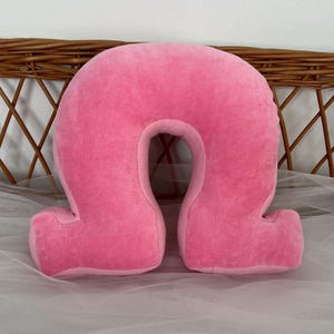 May include: A plush, pink travel pillow in a U-shape. The pillow is made of a soft, velvety material and has a curved design to support the neck. The pillow has two small, rectangular extensions at the bottom.