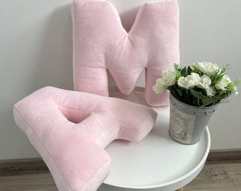 Könnte beinhalten: Zwei rosa Plüsch-Buchstabenkissen, ein 'M' und ein 'A', sind auf einem weißen Tisch mit einer kleinen Topfpflanze mit weißen Blüten angeordnet.