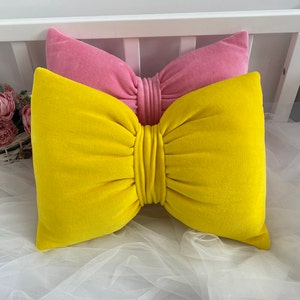 Puede incluir: Dos almohadas decorativas con diseño de lazo. La almohada superior es rosa y la almohada inferior es amarilla.