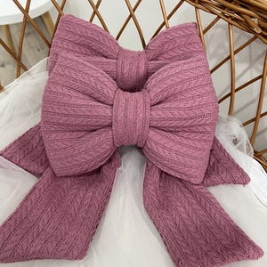 May include: A large, mauve bow made from textured fabric. The bow is centrally positioned with long, flowing tails. The fabric has a subtle, woven pattern. The bow rests on white, sheer fabric, with a wicker chair in the background.