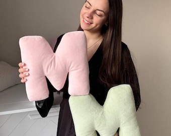 May include: Two plush letter pillows, one pink and one green, in the shape of the letter M. A person is holding the pillows.