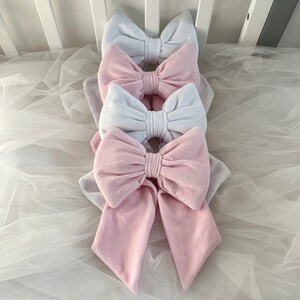 May include: Four fabric bows stacked in a pyramid shape, two white and two pink. The bows are made from a soft, plush fabric and have a satin ribbon tied around the centre.