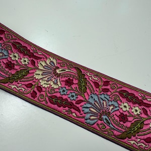 Könnte beinhalten: Ein gewebtes Stoffband mit einem leuchtend pinkfarbenen Hintergrund und einem komplexen Blumenmuster. Das Design zeigt stilisierte Blüten und Blätter in Hellblau, Creme, Dunkelrot und Olivgrün, abgerundet durch dunkel gefärbte Kanten.