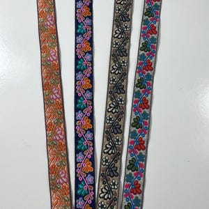 Op de afbeelding: Vier verticale stroken decoratief stoffen lint met ingewikkeld bloemenborduurwerk. De eerste is oranje met roze en groene bloemen, de tweede marineblauw met veelkleurige bloesems, de derde metallic goud met zwarte bloemmotieven en de vierde lichtgrijs met rode en blauwe bloemmotieven.