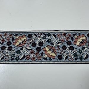 Könnte beinhalten: Ein breites Stoffband mit hellblauem Hintergrund und einem komplexen gestickten Blumenmuster. Das Design umfasst pfirsich- und cremefarbene Blüten, dunkelbraune runde Beeren und kleine grüne Blätter, alles eingefasst von einem dunklen Rand.