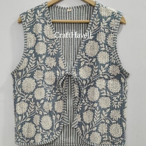 May include: A sleeveless, quilted vest with a floral pattern in white on a blue-grey background. The vest has a striped interior and a tie closure. The vest is hanging on a wooden hanger.
