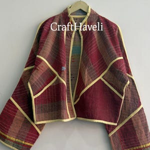 May include: A cropped jacket featuring a maroon, brown, and yellow plaid pattern with visible white running stitch details. The garment has long sleeves, a stand collar, and light yellow trim along the edges. The text CraftHaveli is visible.
