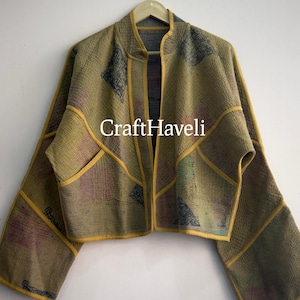 Puede incluir: Una chaqueta corta de mangas largas y anchas con cuello mao, que presenta una base de color verde oliva apagado con sutiles patrones multicolores y ribetes amarillos a lo largo de las costuras y bordes. El texto CraftHaveli está impreso en el centro.
