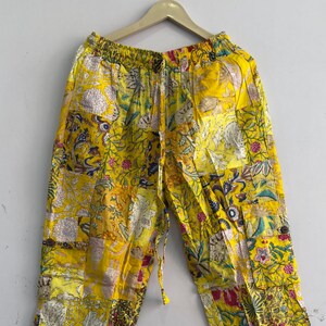 Op de afbeelding: Gele broek met een patchworkontwerp met diverse bloemen- en botanische patronen in tinten rood, blauw en wit. De broek heeft een elastische tailleband met een trekkoord en wordt getoond op een houten kledinghanger.