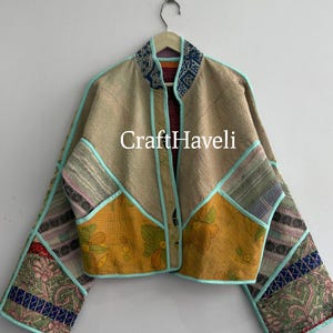May include: A cropped jacket featuring a patchwork design of beige, yellow, and multi-colored floral fabric panels. The garment has wide sleeves, a mandarin collar, and light blue piping along the edges. The text CraftHaveli is centered.