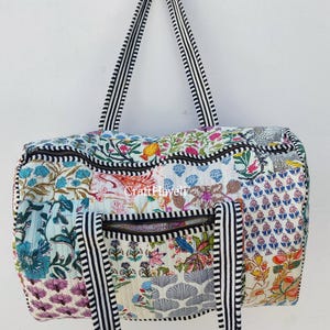 May include: A white quilted duffel bag with black and white striped straps. The bag is decorated with a patchwork of colourful floral prints. The bag has a zipper closure and a top handle.