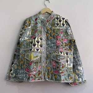 Könnte beinhalten: Eine gesteppte Jacke in hellblauer Grundfarbe mit einem mehrfarbigen Blumenmuster in Rosa, Gelb und Grün. Das Kleidungsstück hat einen Stehkragen, zwei Vordertaschen mit Klappen und durchgehend schwarz-weiß gestreifte Kantenabschlüsse.