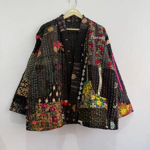 May include: A black quilted jacket featuring a patchwork design with various floral and geometric patterns in red, yellow, green, and white. The garment has long sleeves, an open front, and visible white running stitch details throughout. Text reads CraftHaveli.