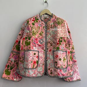 May include: A quilted jacket in a light pink hue featuring a vibrant floral and botanical print in shades of green, red, and white. The garment has a round neckline, long sleeves, two front patch pockets, and black and white striped trim.