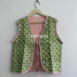 May include: A sleeveless quilted vest featuring a vibrant green base with a repeating pink and blue floral pattern. The garment has a V-neckline, a front tie closure, and a contrasting red and white striped interior lining and trim. Text reads CraftHaveli.