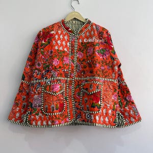 以下が含まれることがあります： 鮮やかなオレンジ色のキルティングジャケットで、多色の花柄とペイズリー柄のブロックプリントが施されています。マンダリンカラー、長袖、2つのフロントパッチポケット、黒と白のストライプのトリムが特徴です。テキストにはCraftHaveliと書かれています。