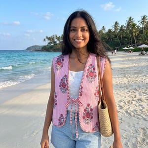 May include: A sleeveless pink quilted vest featuring a white vertical pinstripe pattern and a floral print in shades of red and blue. The vest has a rounded hem, a front tie closure, and decorative stitching along the edges, worn over a white top.