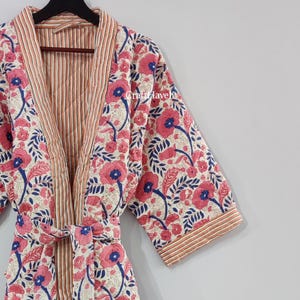 Może przedstawiać: Biała bawełniana szlafrok z kwiatowym nadrukiem w odcieniach różowego i niebieskiego. Szlafrok ma pasek do wiązania i podszewkę w paski.