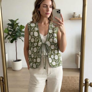 Könnte beinhalten: Eine ärmellose Weste mit einem grünen und weißen floralen Blockdruckmuster. Die Weste hat einen V-Ausschnitt, einen vorderen Verschluss zum Binden mit dünnen Stoffbändern und einen gesäumten Rand mit sichtbaren Ziernähten am Kragen.
