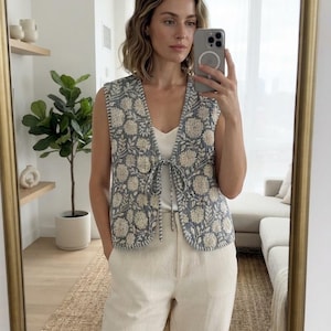 Puede incluir: Un chaleco sin mangas con un estampado floral en azul y blanco y textura acolchada. El chaleco presenta un escote en V, un cierre frontal de lazo con cordones de tela a juego y detalles de costura visible a lo largo de los bordes.