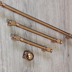 Pode incluir: Quatro puxadores de armário em ouro rosa com base quadrada e pomo redondo na extremidade. Os puxadores estão dispostos em forma de escada sobre uma superfície de madeira.