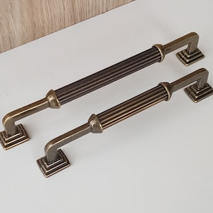 May include: Two antique gold cabinet door pulls with a decorative, fluted design. The pulls have a square base with a raised edge.