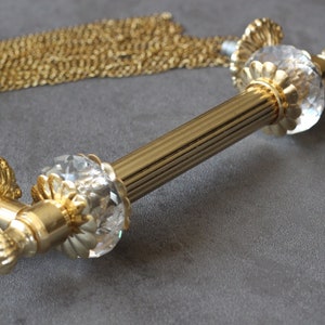 Puede incluir: Pomo de puerta dorado con terminales de cristal. El pomo tiene un diseño acanalado y está unido a una cadena.