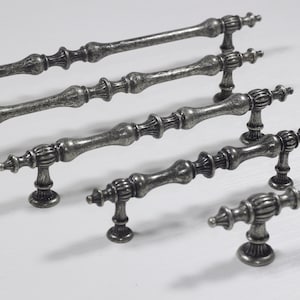 以下が含まれることがあります： さまざまなサイズとスタイルのアンティークシルバーの引き出しノブが5つあります。ノブは装飾的なデザインで、金属製です。