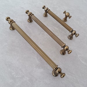 Peut inclure: Ensemble de quincaillerie d'armoire en laiton antique. L'ensemble comprend trois longues poignées et deux petits boutons. Les poignées ont un design nervuré et des extrémités arrondies. Les boutons ont un design similaire. La quincaillerie est en métal et a un aspect vintage.