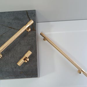 Puede incluir: Tres tiradores de armario de color dorado. Dos son largos y rectos, y uno es más corto y horizontal.