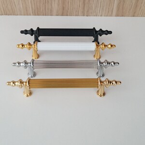 May include: Four cabinet pulls in different finishes: black, white, silver, and gold. Each pull has a fluted design and a decorative finial.