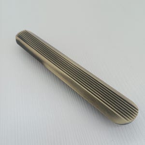 May include: A long, narrow, brushed metal cabinet pull with a grooved design. The pull is a light gold color.