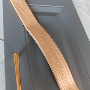 Può includere: Una maniglia per mobili in legno di rovere chiaro con un design curvo è posizionata su un'anta grigia. È visibile anche una maniglia in metallo dorato. La venatura del legno è evidente, suggerendo un design moderno per cucina o mobili.