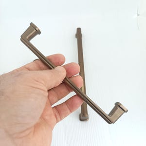 May include: Bronze-colored cabinet pulls with a rectangular design. The pulls have a straight bar with a square end on each side. One pull is held in a hand, and another is in the background.