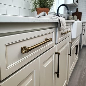 Puede incluir: Primer plano de gabinetes de cocina con herrajes de latón antiguo. Los gabinetes de color gris claro presentan paneles rectangulares. Los herrajes incluyen tiradores largos y tiradores verticales más pequeños, todos en un acabado de latón cálido.