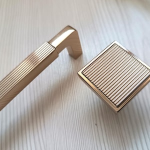 May include: A gold-colored cabinet pull and knob set. The pull is rectangular with a ridged design, and the knob is square with a matching ridged pattern. The hardware is made of metal and has a shiny finish. Ideal for kitchen or bathroom cabinets.