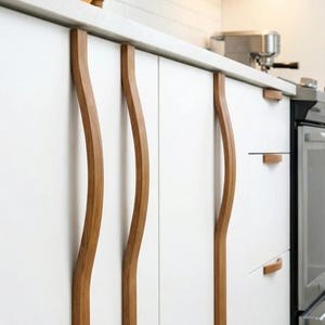 May include: White kitchen cabinets with curved wooden handles. The handles have a natural wood grain finish and are vertically oriented. The cabinets are part of a modern kitchen design, with a white countertop and stainless steel appliances.