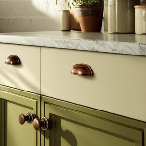 Może przedstawiać: Zbliżenie na szafki kuchenne z antycznym, brązowym osprzętem. Górne szafki są w kolorze jasnego kremu, a dolne w kolorze oliwkowej zieleni. Blat jest z białego marmuru. Okucia obejmują uchwyty w kształcie filiżanek i okrągłe gałki.