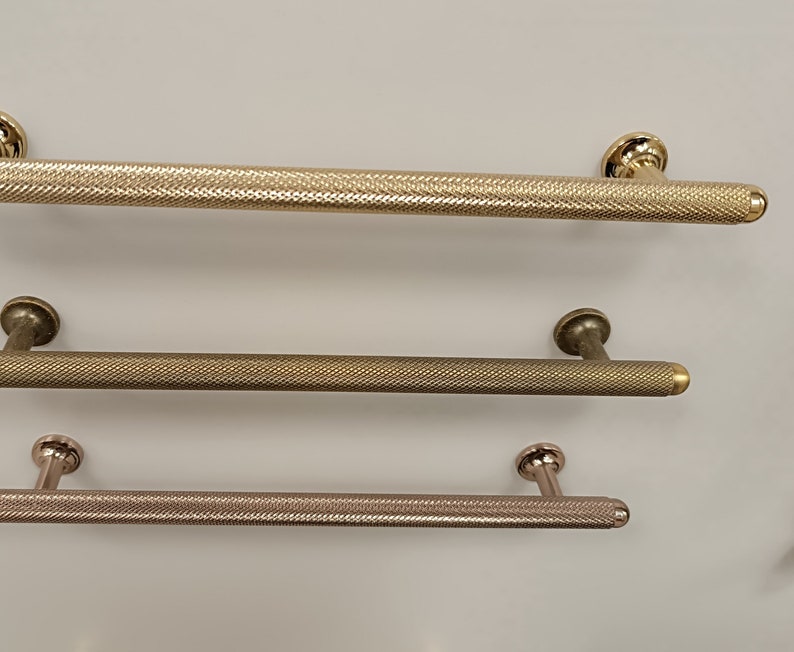 May include: Three different metal cabinet door pulls with a textured finish. The top pull is gold, the middle pull is bronze, and the bottom pull is a light rose gold.