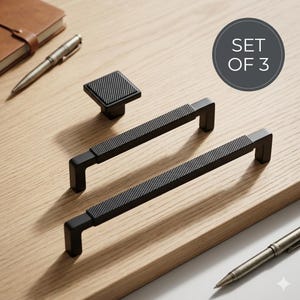 May include: A set of three matte black cabinet hardware pieces, including one square knob and two rectangular drawer pulls. Each piece features a textured, knurled center section. A circular graphic in the corner displays the text SET OF 3.